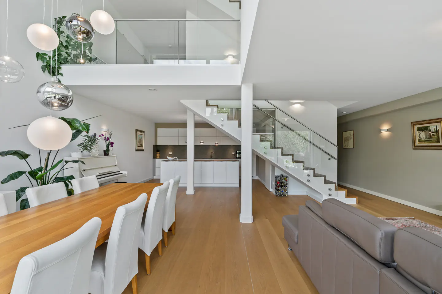 Modern open-plan home with a wooden dining table, white chairs, a sleek kitchen, and a glass-railed staircase.