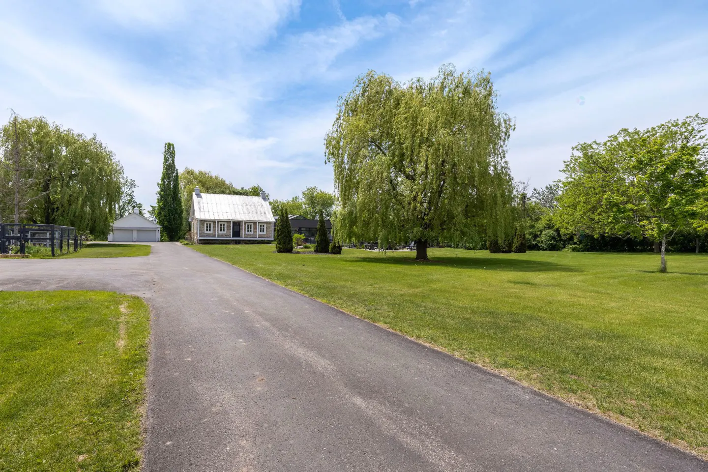 A long asphalt driveway leads to a charming white cottage with a metal roof, surrounded by lush green lawns and large trees.