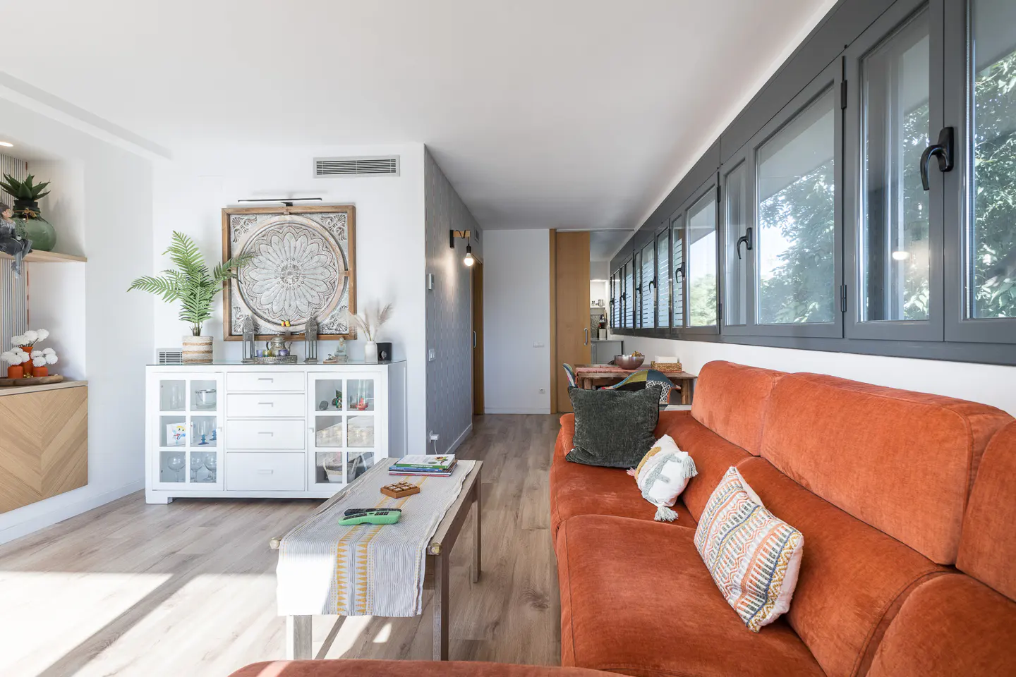 Bright living room with orange sofa, wood floors, and white cabinet. Gray framed windows line the wall above the sofa. A coffee table sits in front.