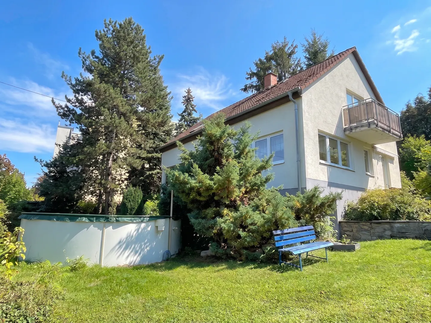 A two-story house with a balcony, a blue bench, and a pool in the backyard on a sunny day.