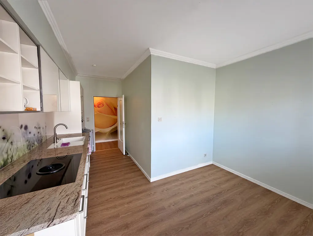A bright kitchen with wood floors, light green walls, and white cabinets. A doorway shows a rose mural in the next room.