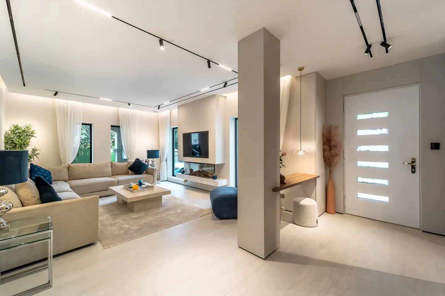 A modern living room with a beige sectional sofa, a TV over a fireplace, and a white front door.