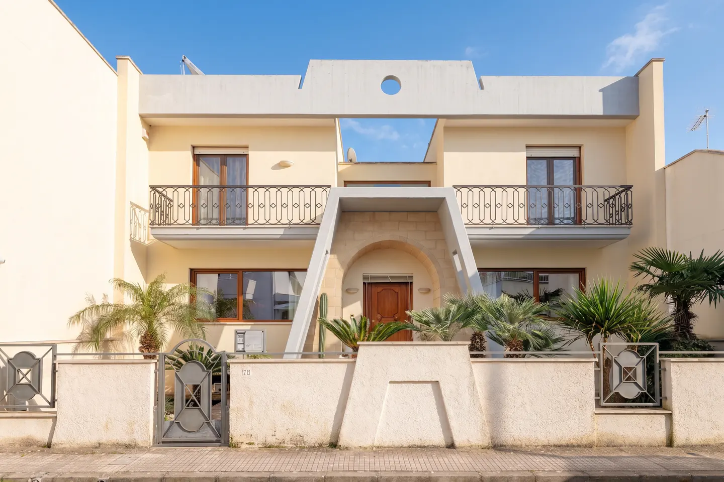 Two-story cream house with balconies, a brown door, and a unique architectural design under a clear blue sky.