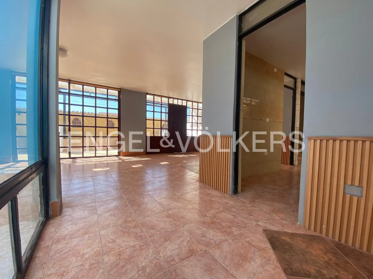 A spacious room with terracotta tile flooring, large windows, and gray walls. A doorway leads to another room. The Engel & Völkers logo is visible.
