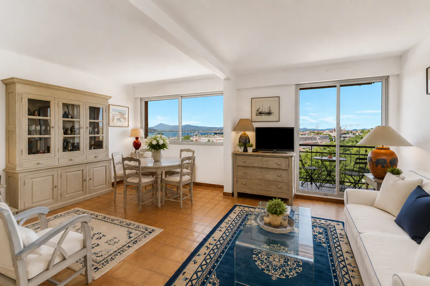 Bright living room with dining area, white sofa, blue rug, and balcony doors overlooking a scenic bay and mountain view.
