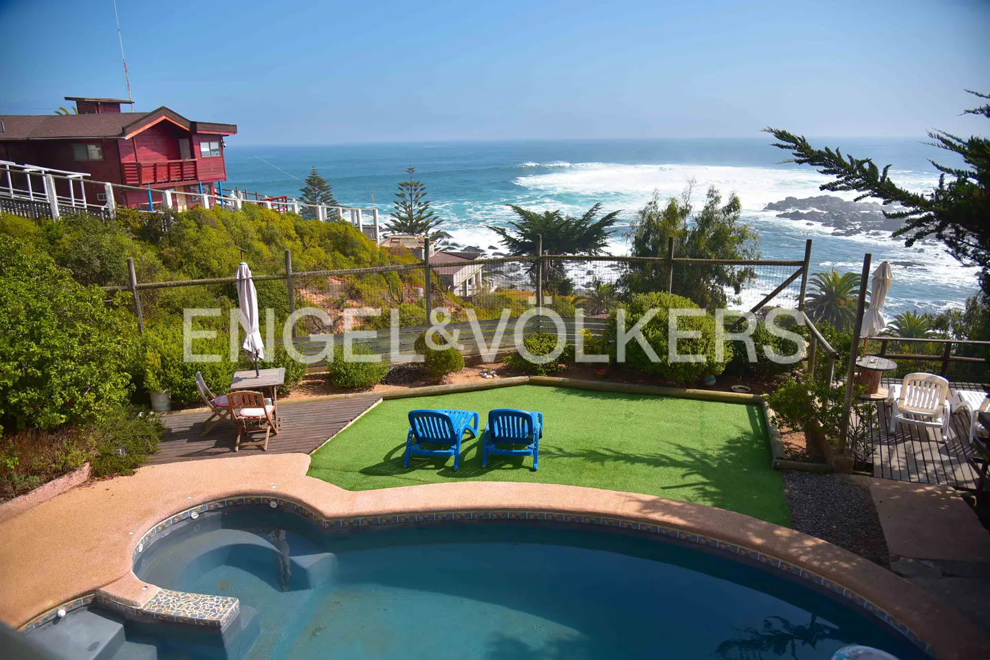 A backyard with a pool, green lawn, and two blue lounge chairs overlooking the ocean. A red house sits on a hill in the background.