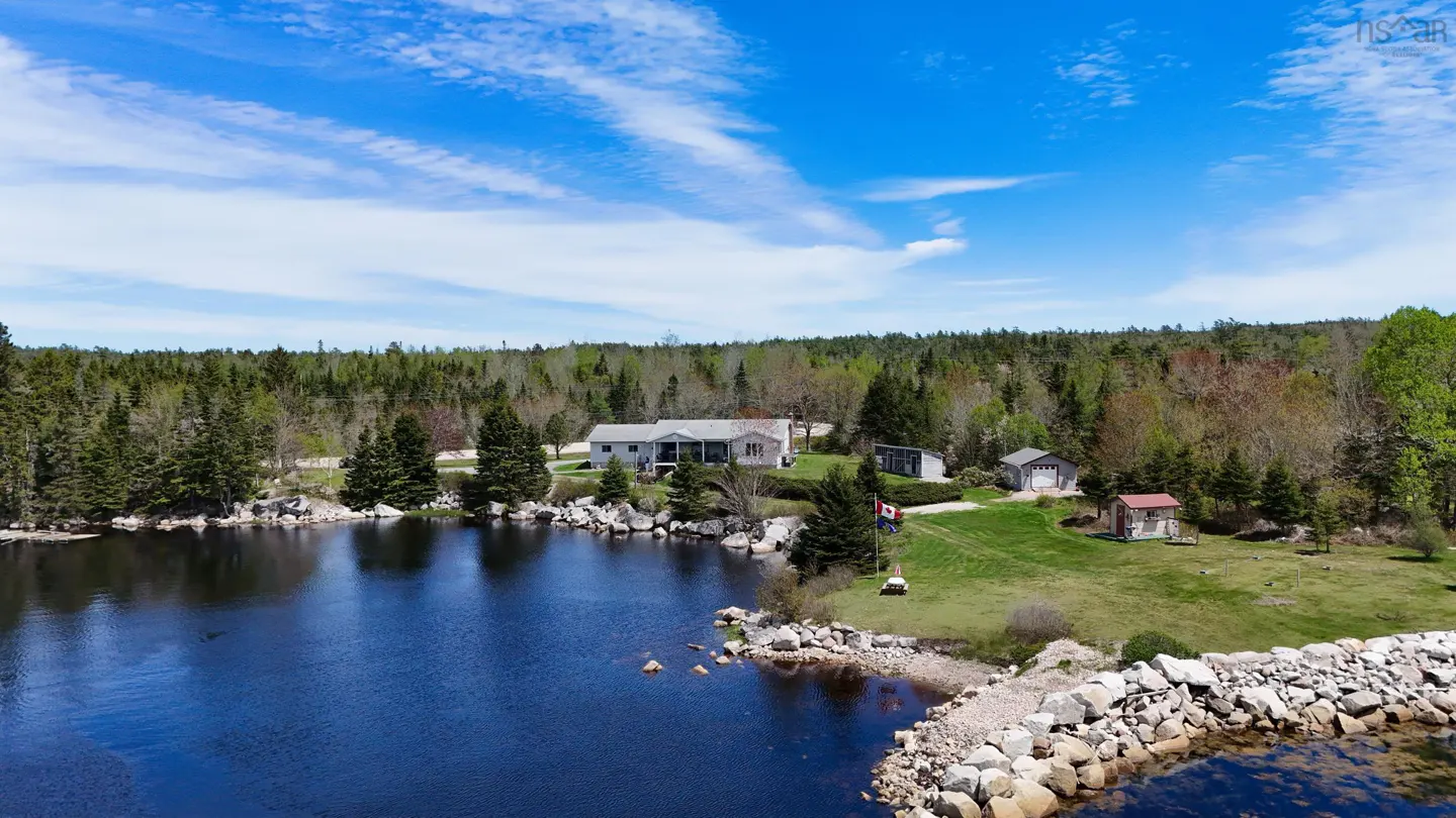 Waterfront property with a gray house, green lawn, and forest backdrop under a blue sky with scattered clouds.