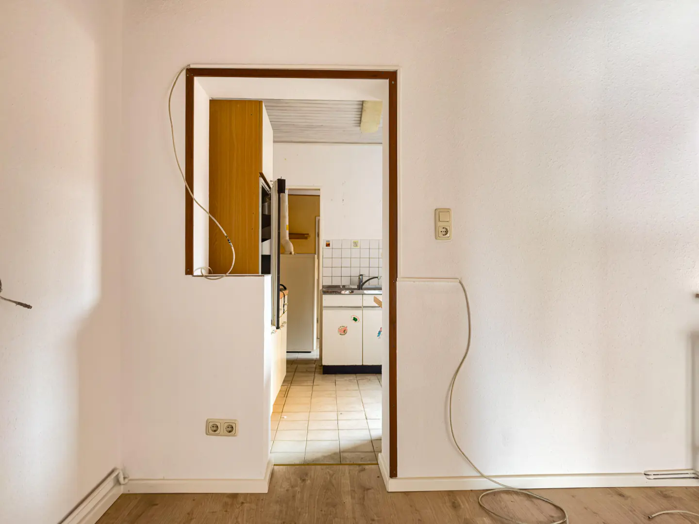 A view of a kitchen through a doorway. The walls are white, and the floor is wood. The kitchen has white cabinets and a sink.