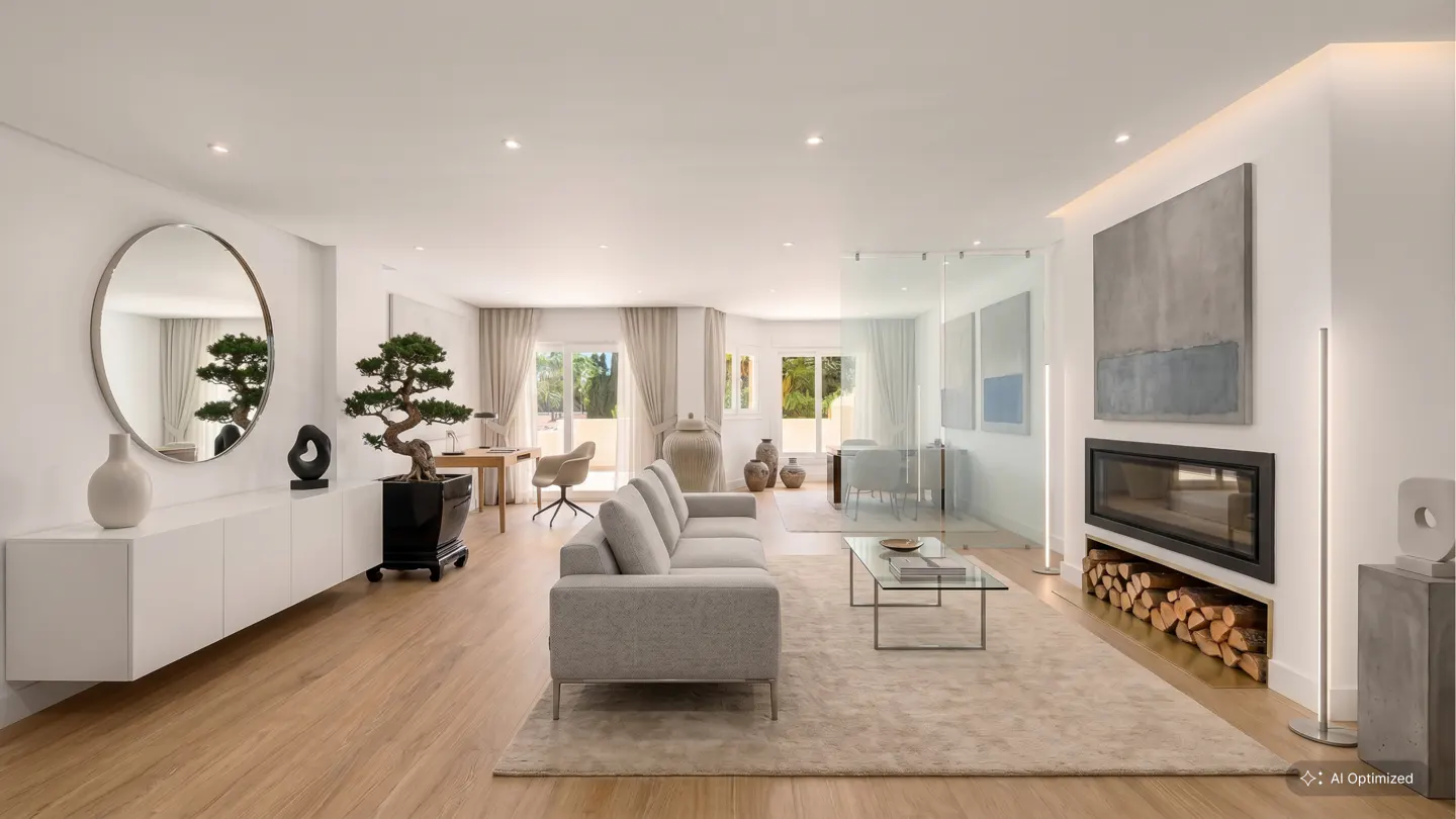 Bright, modern living room with a gray sofa, glass coffee table, and fireplace with stacked wood. A bonsai tree sits near a round mirror.