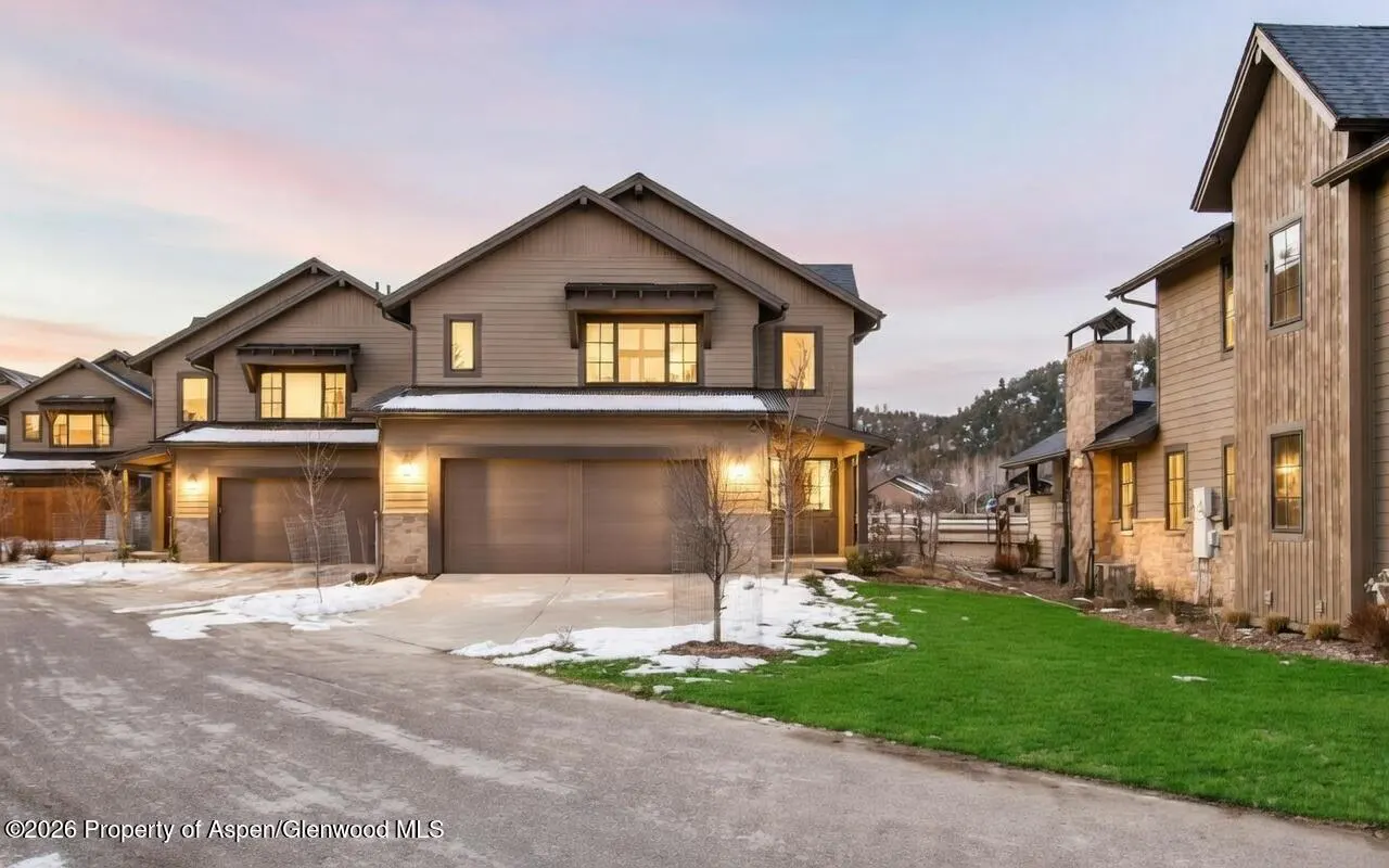 Exterior view of modern, two-story townhouses with attached garages, green lawns, and snow patches at dusk.