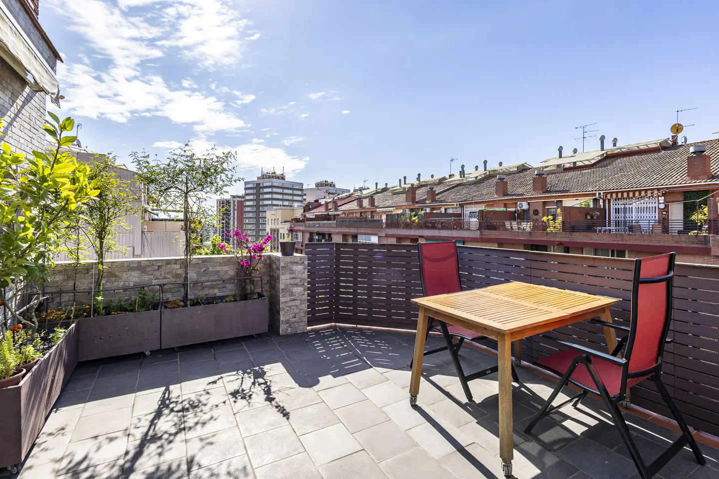 Sunny rooftop terrace with a wooden table, two red chairs, potted plants, and a view of surrounding city buildings.