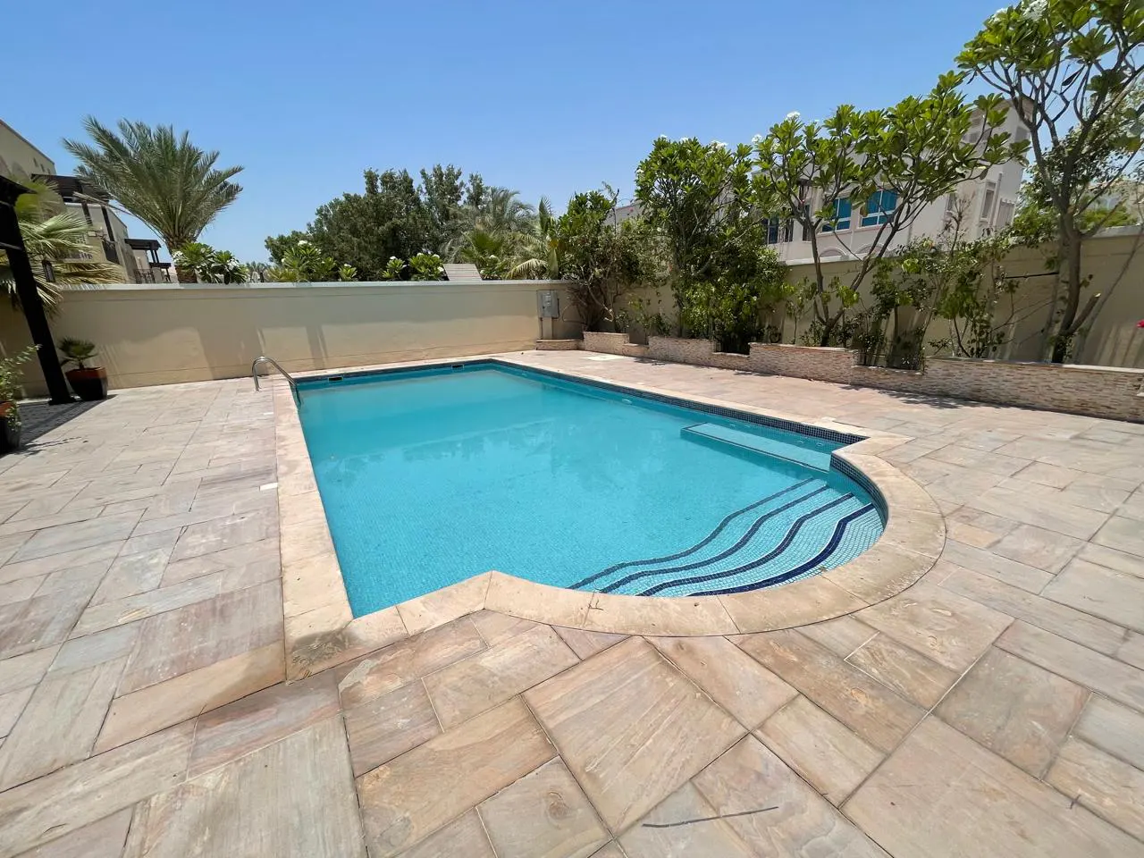 A backyard swimming pool with blue water, surrounded by stone pavers and green trees under a blue sky.