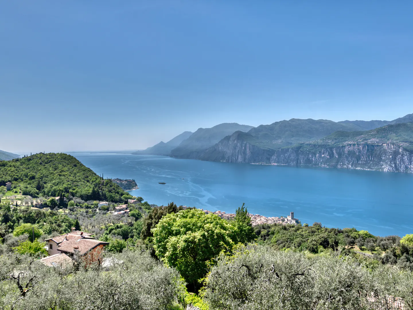 Scenic view of Lake Garda, Italy, with blue water, green hills, mountains, and a small town with a castle.