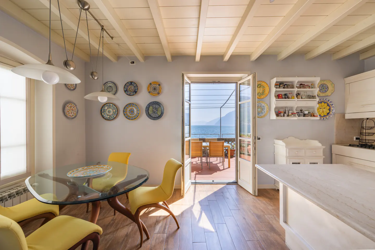 Bright kitchen with yellow chairs, a glass table, and decorative plates on the wall. Open doors lead to a patio with a view of the sea.