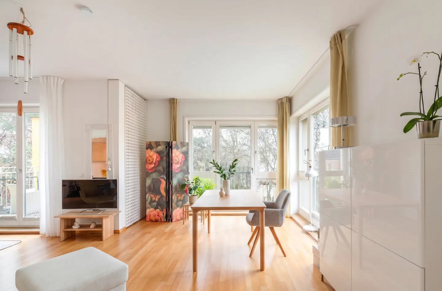 Bright living room with wood floors, white walls, and large windows. A dining table and chairs sit near a floral screen. TV and ottoman are visible.