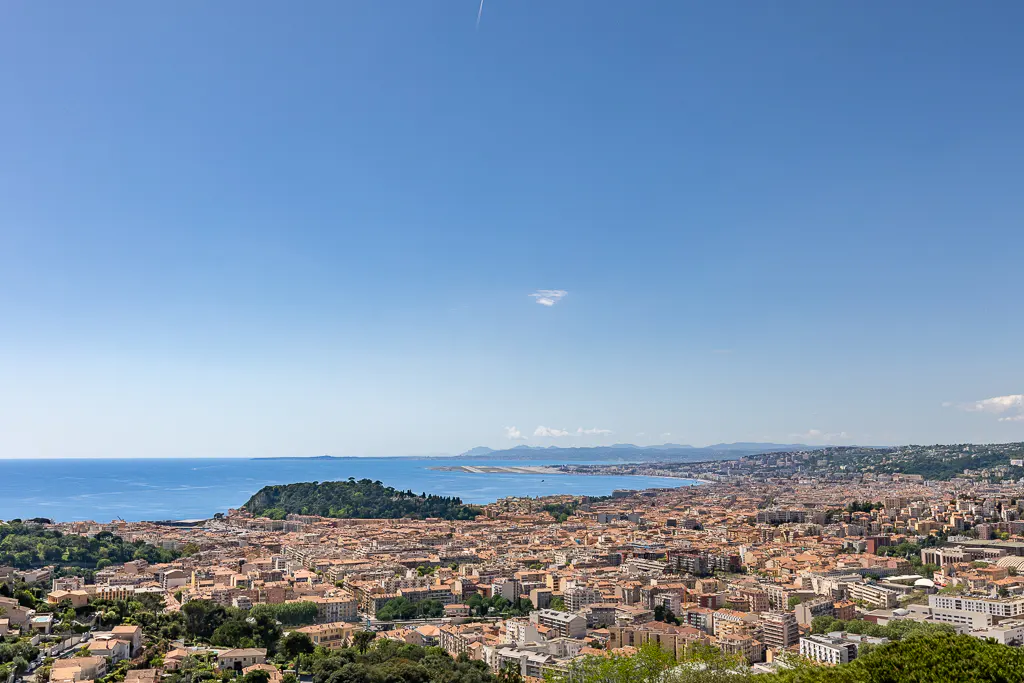 Aerial view of Nice, France, showing the city's buildings with orange roofs, the blue Mediterranean Sea, and a clear blue sky.