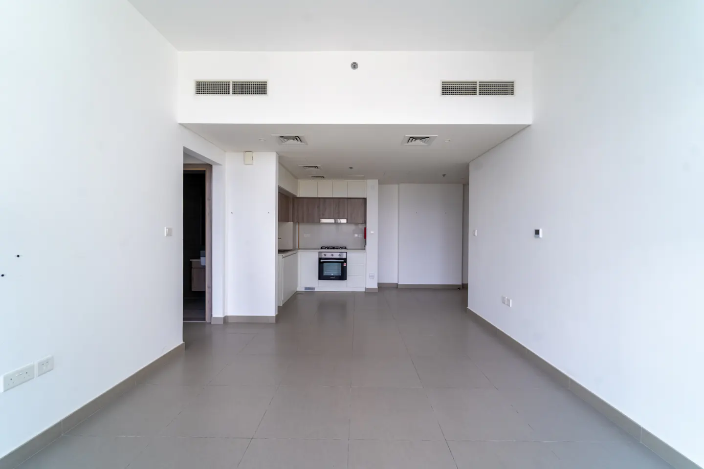 An empty apartment with white walls and gray tile floors. A kitchen is visible in the background.
