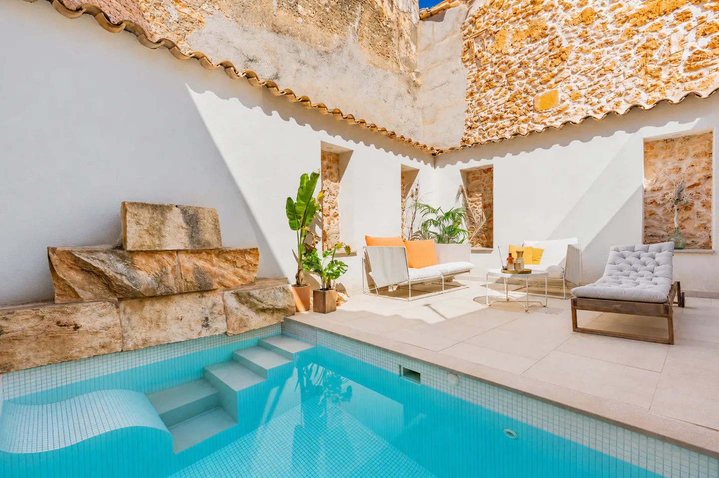 Outdoor courtyard with a turquoise pool, stone steps, and white lounge furniture with orange pillows. Plants add greenery. Walls are white and stone.