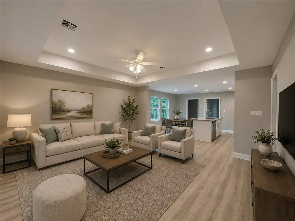 Open-concept living room with beige sofa, two armchairs, and a wooden coffee table on a neutral rug. Kitchen and dining area in background.