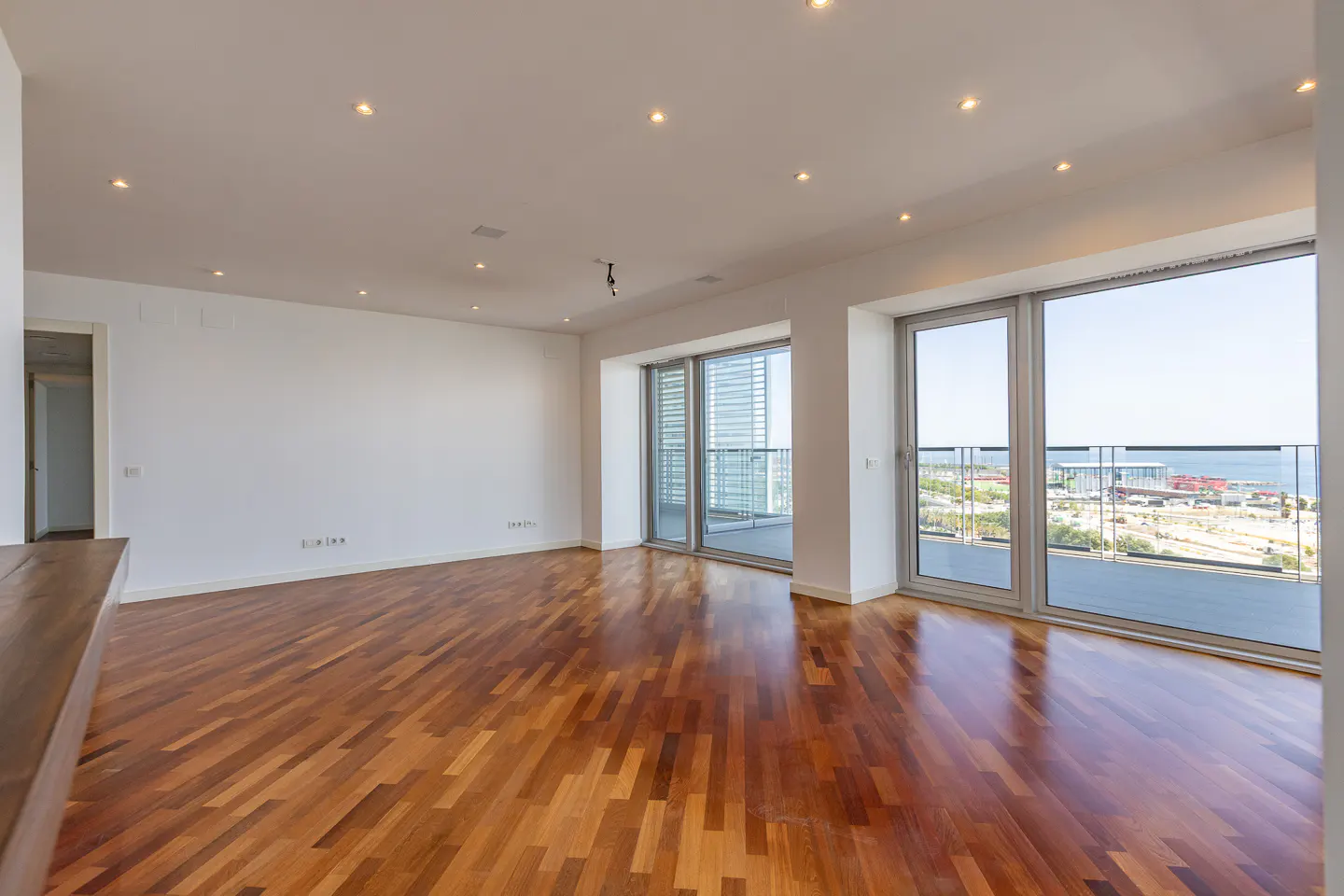 Bright, empty living room with polished hardwood floors, white walls, recessed lighting, and floor-to-ceiling sea views.