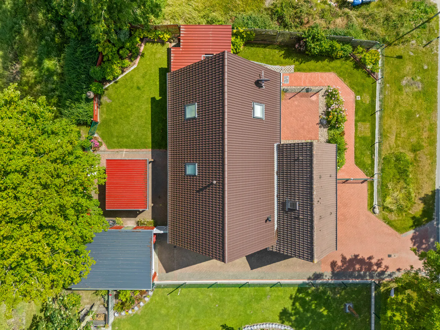 Aerial view of a brown-roofed house with green lawn, red brick driveway, and surrounding trees.