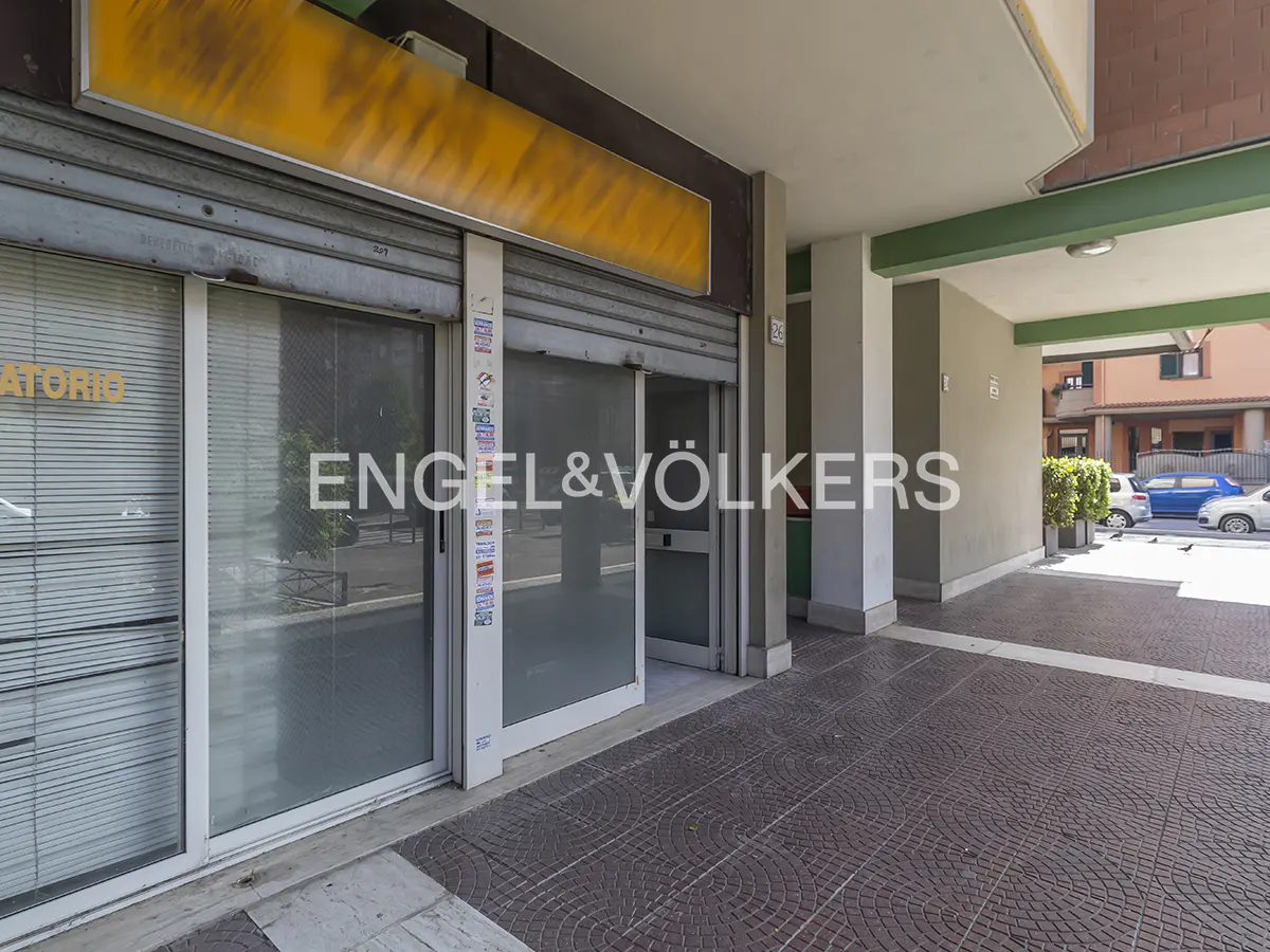 Exterior view of a commercial space with Engel & Völkers logo on the glass door. The building has a covered walkway with gray columns and a brick-patterned floor.