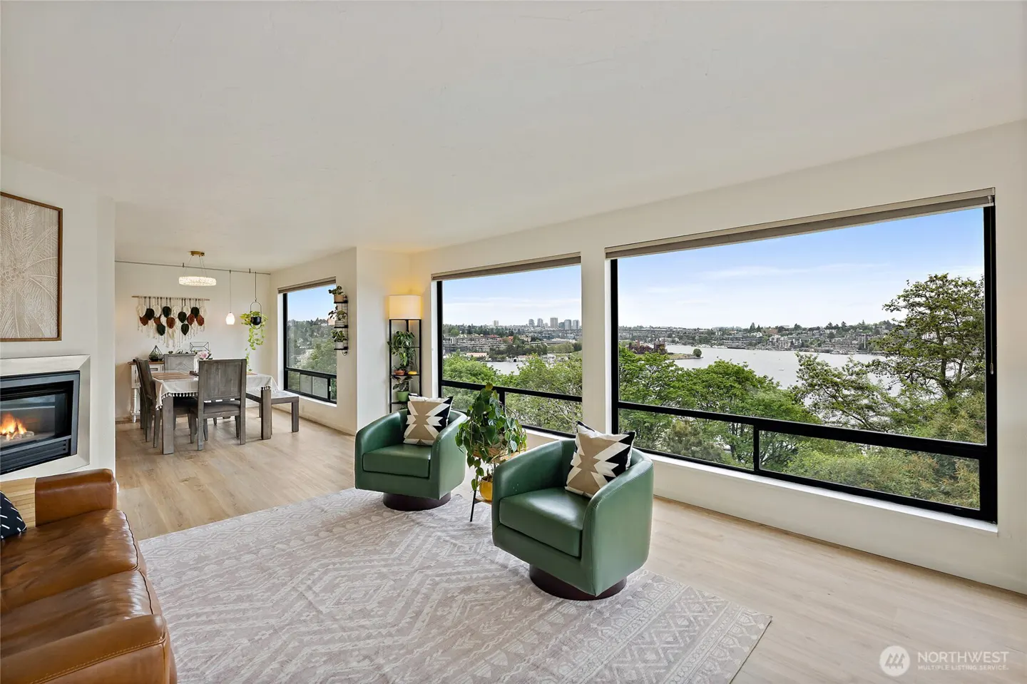 Bright living room with a fireplace, dining area, and large windows overlooking a lake and city skyline. Two green chairs sit on a patterned rug.