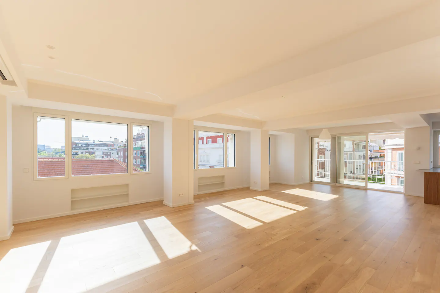 Bright, empty room with light wood floors, white walls, and large windows showing city views. Balcony access visible.