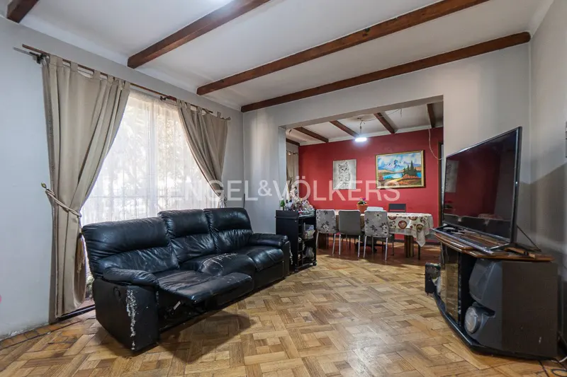 Living room with a black leather sofa, wooden floors, and a dining area with a red wall in the background. Curtains frame a window.