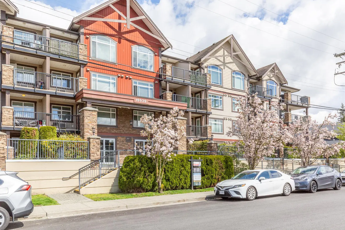 Exterior view of a three-story apartment building with balconies, flowering trees, and parked cars.