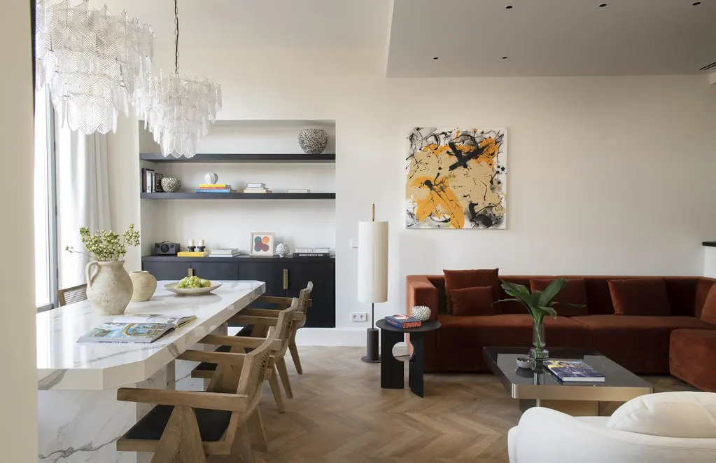Bright, modern living space with a marble island, wooden chairs, and a rust-colored sofa. Abstract art hangs above the sofa.