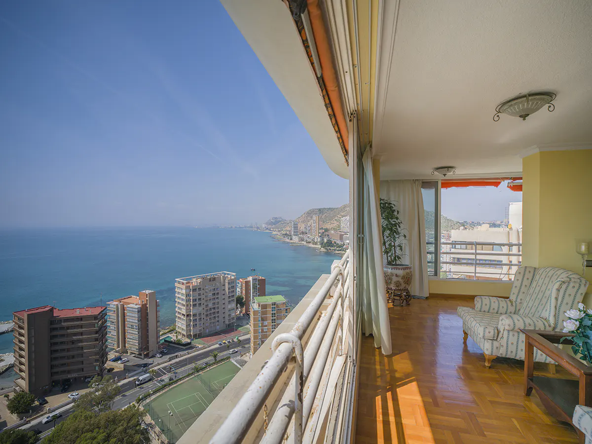 Balcony view of the ocean and city. Inside, a patterned armchair sits on a wood floor.