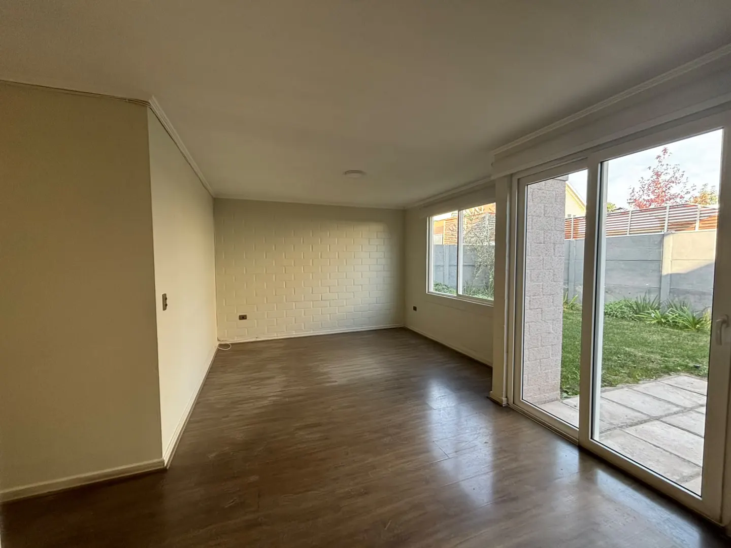 Empty living room with light-colored brick walls, dark wood-look flooring, and large sliding glass doors to a backyard.