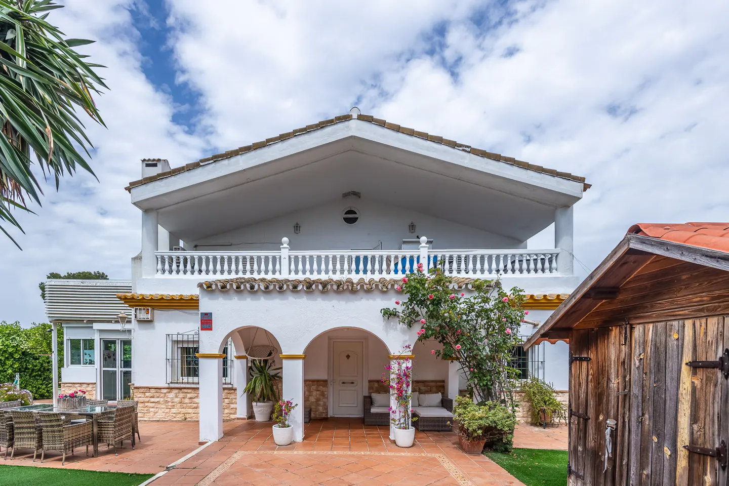 Two-story white house with arched porch, red tile patio, and outdoor furniture under a cloudy sky.