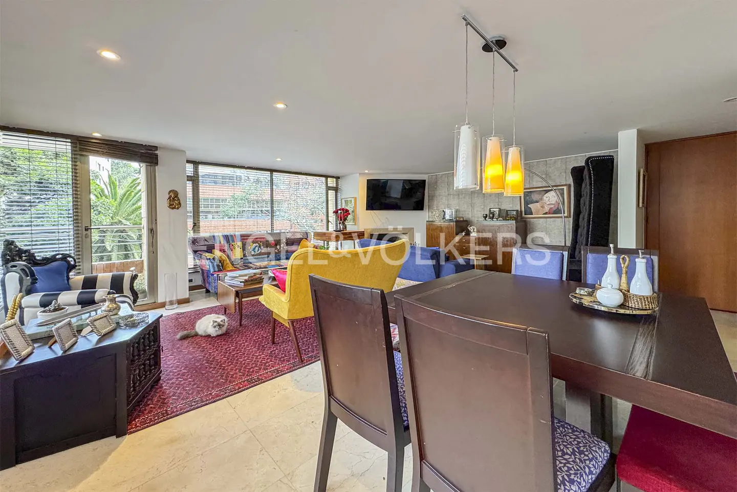 A bright living and dining area with a dark wood table, colorful furniture, and a cat on a red rug.
