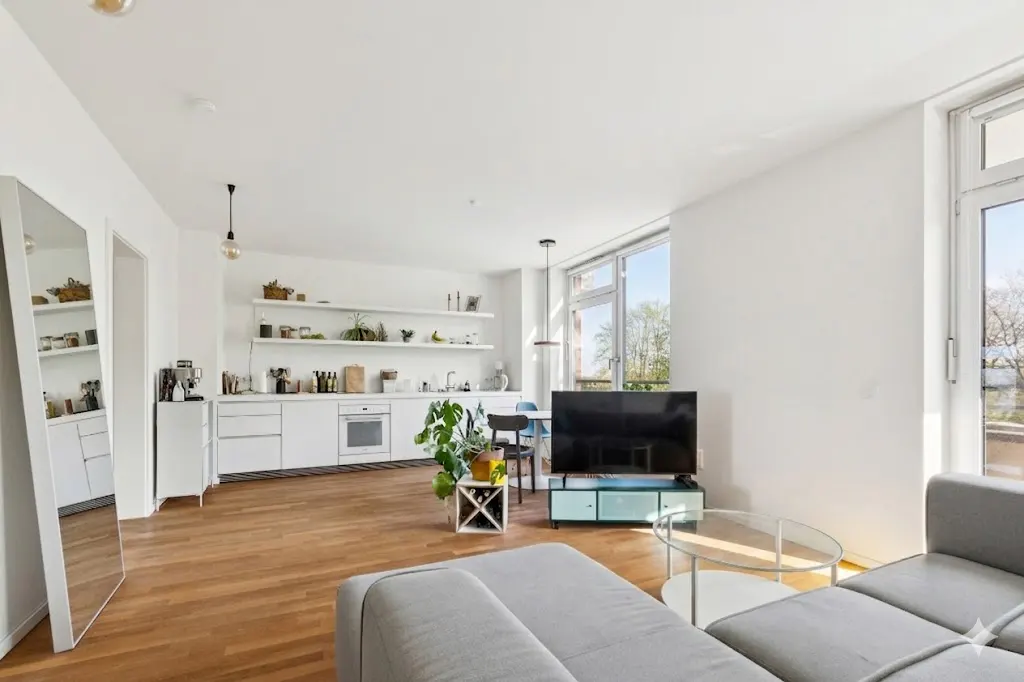 Bright, open-concept apartment with white walls, wood floors, and a gray sectional sofa. Kitchen with white cabinets and open shelving. Large windows.