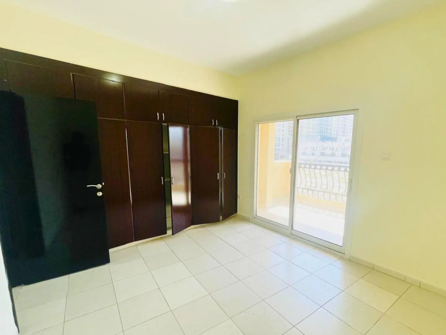 A bright, empty bedroom with cream walls, tiled floor, and dark wood built-in wardrobes. Sliding glass doors lead to a balcony.
