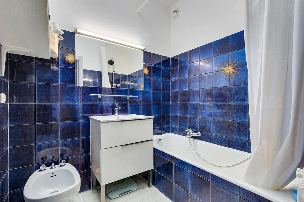 Bathroom with blue tiled walls, white tub with shower curtain, sink vanity, bidet, and mirror.