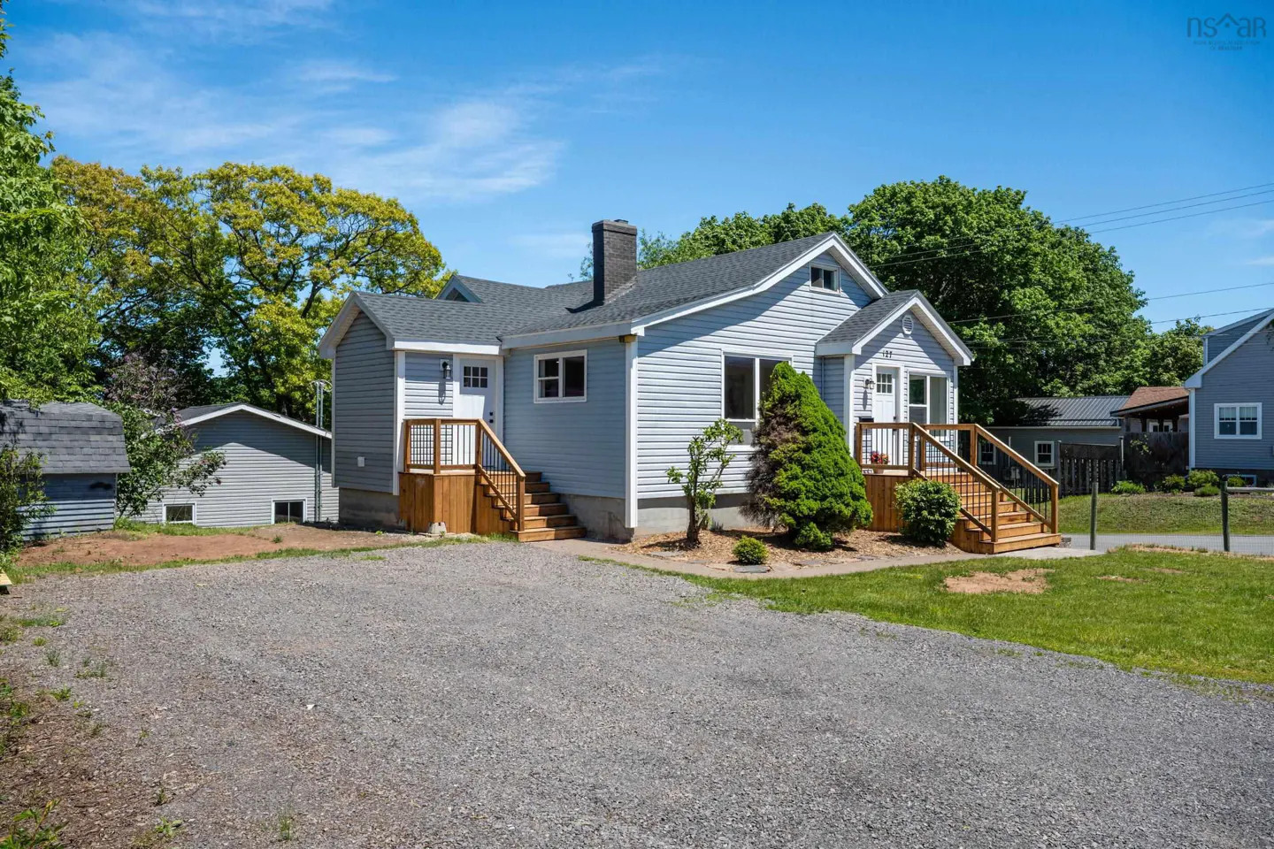Light blue bungalow with a gravel driveway, two wooden entry decks, and a dark chimney under a clear blue sky.