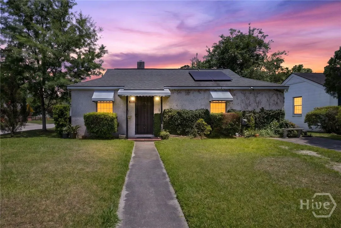 Front exterior of a one-story house with a gray roof, solar panels, and a stone walkway leading to the front door. The sky is a mix of pink and purple.