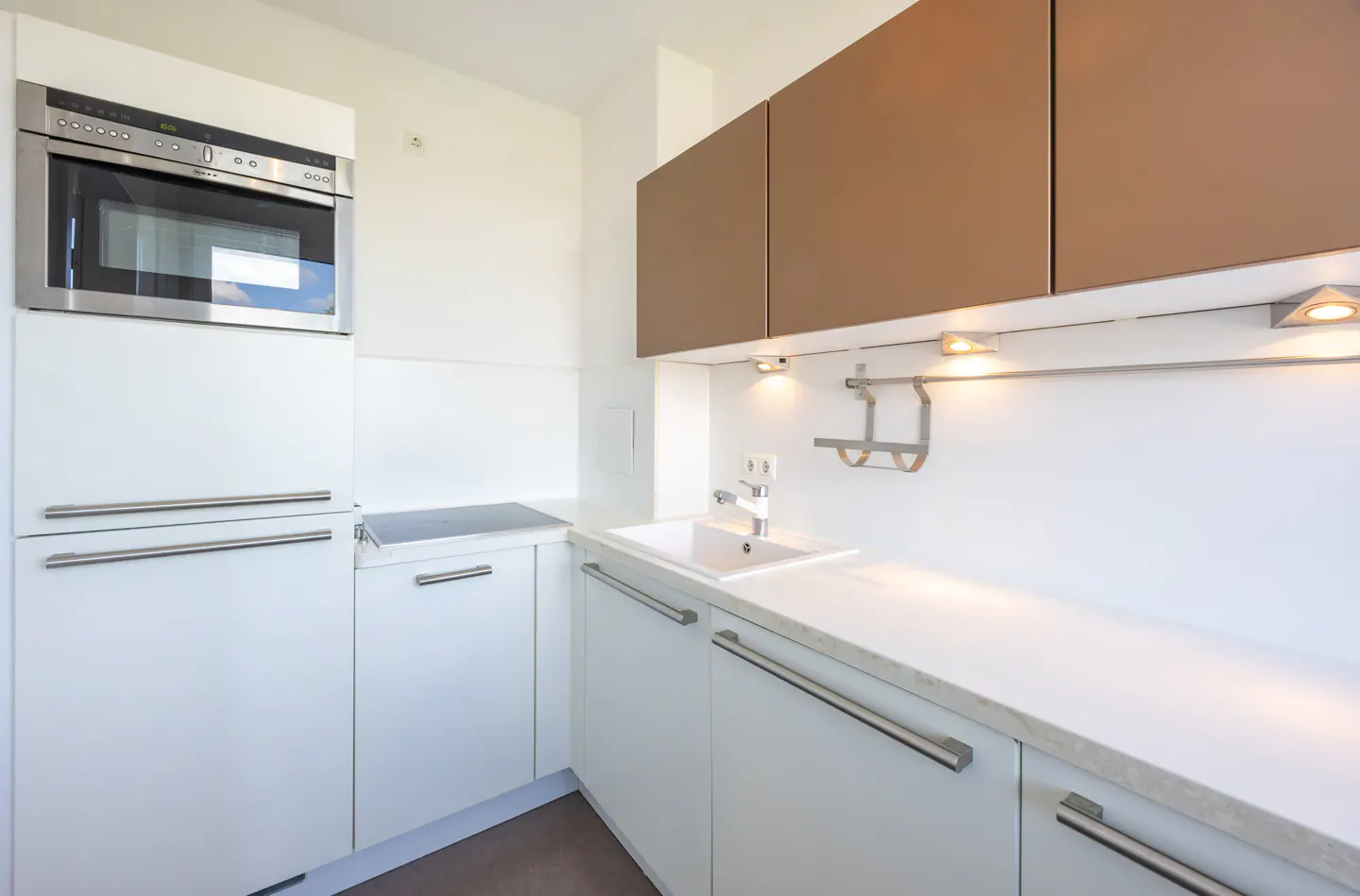 A modern kitchen with white cabinets, stainless steel handles, and a stainless steel microwave. The upper cabinets are brown. A white sink and countertop are visible.