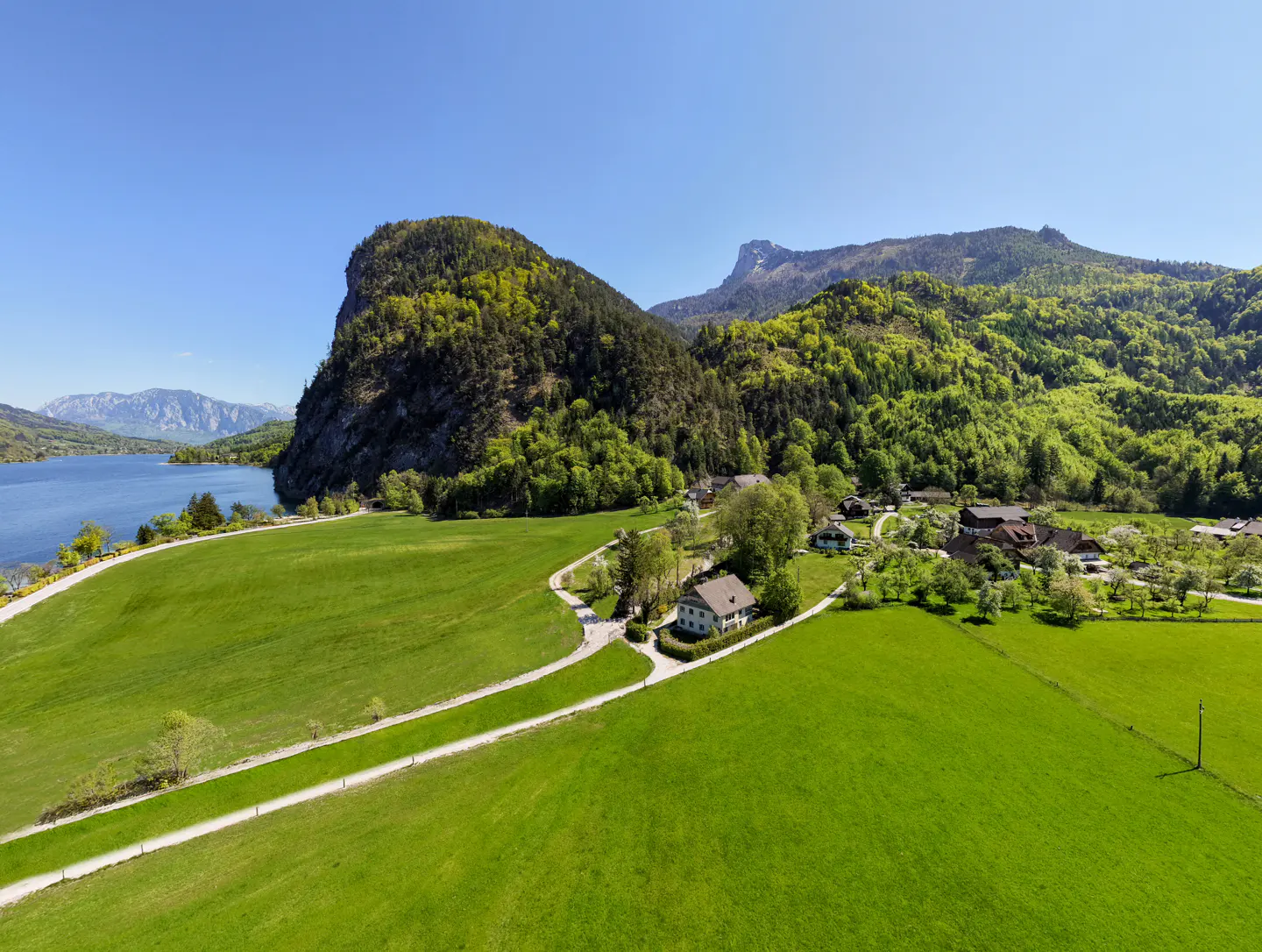 Aerial view of a green meadow, lake, and mountains under a blue sky. A path leads to houses nestled among trees.