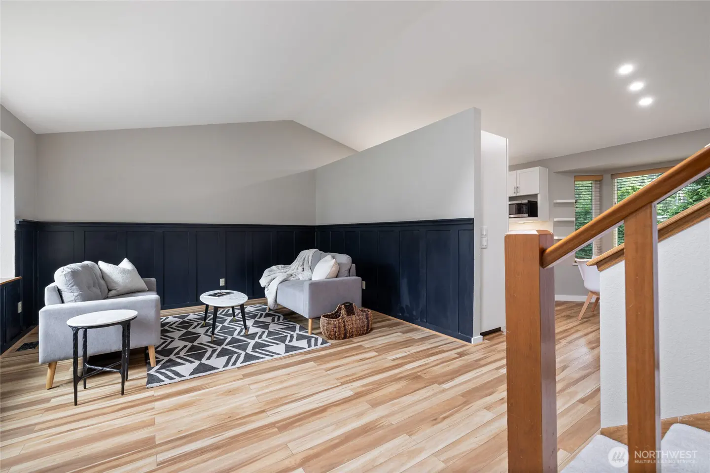 Living room with light wood floors, dark blue wainscoting, two gray armchairs, a geometric rug, and a wooden staircase.