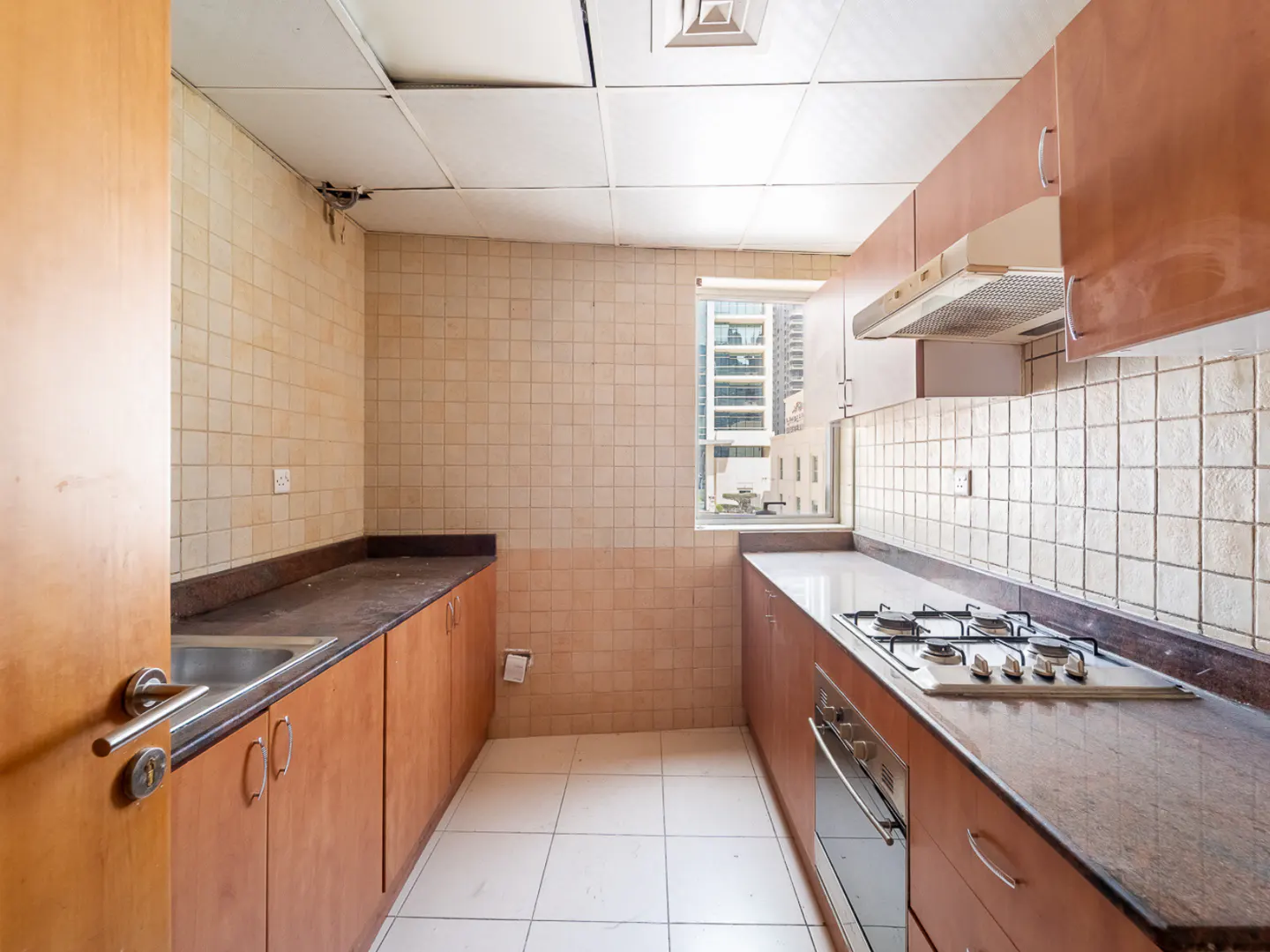 A galley kitchen with brown cabinets, a dark countertop, and a gas stove. A window shows a city view.