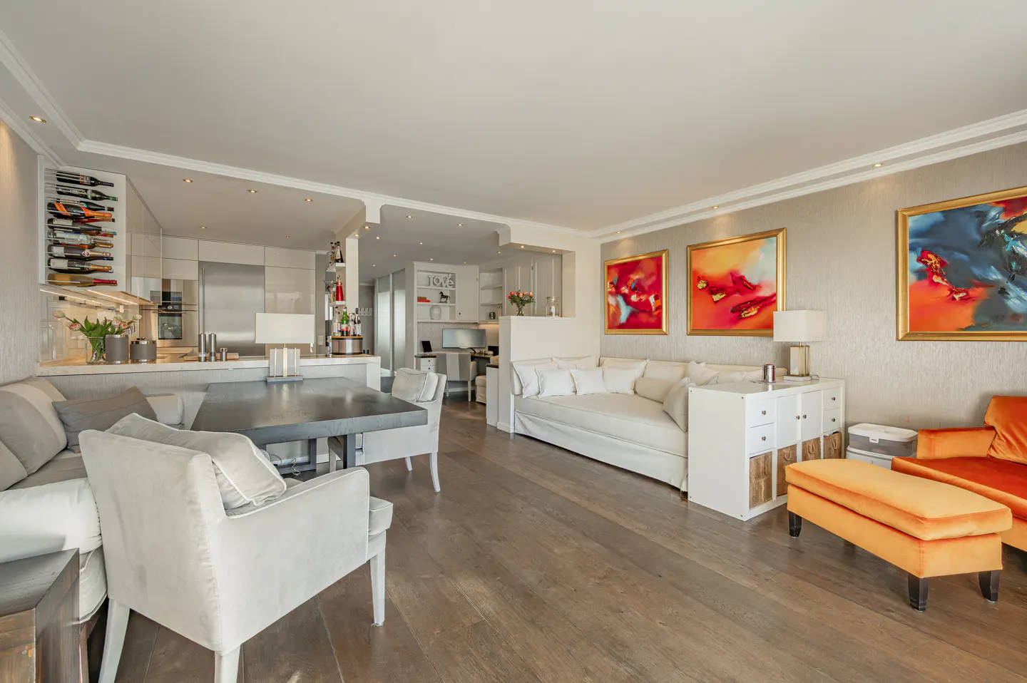 Modern open-plan living room with white sofa, orange armchair, dining area, and vibrant abstract art on beige walls.