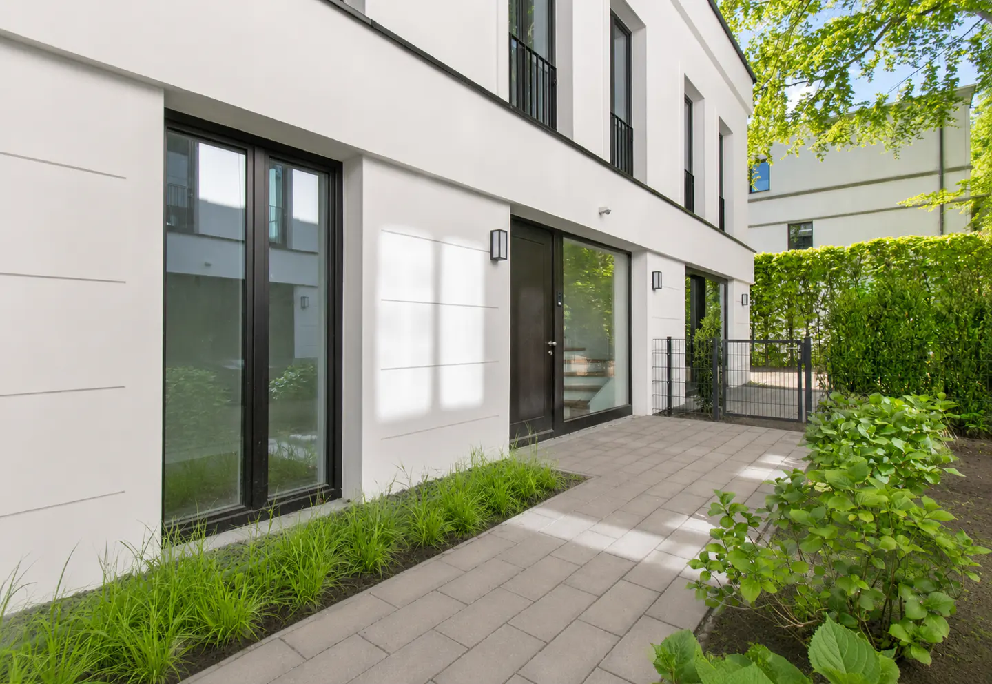 Modern white house with black trim, large windows, and a gray brick walkway. Green plants line the path and a hedge borders the property.