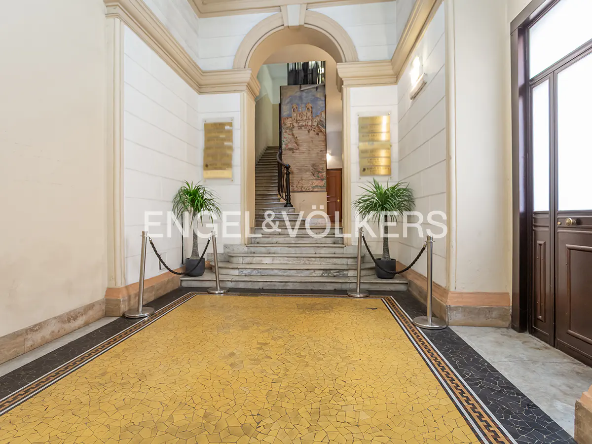 Elegant building entrance with yellow mosaic floor, marble stairs, and potted plants. Archway leads to a staircase with a tapestry.
