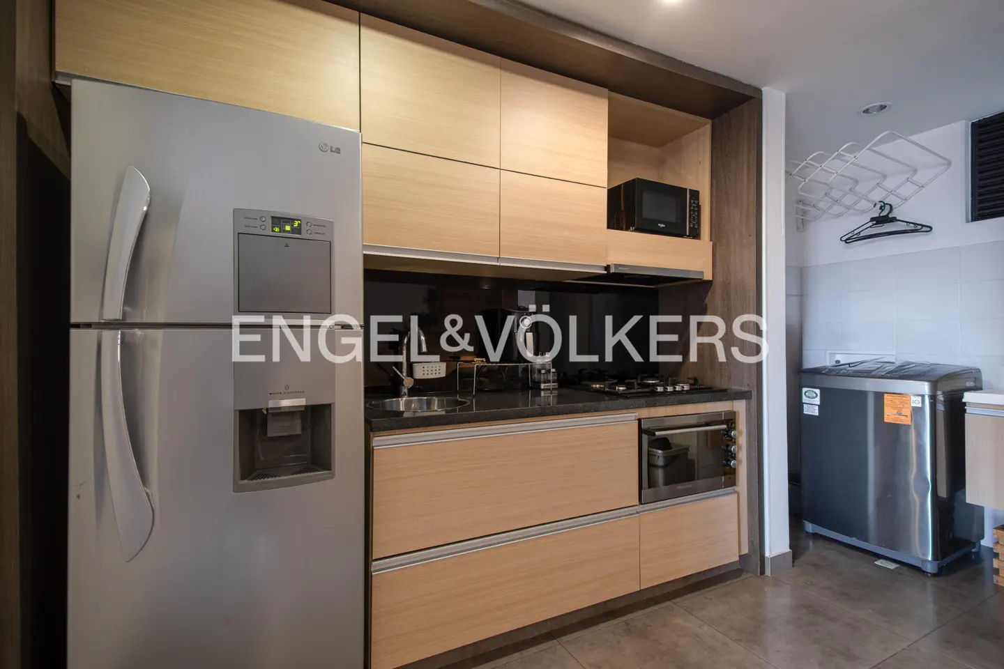 Modern kitchen with stainless steel refrigerator, light wood cabinets, black countertop, microwave, and washing machine.