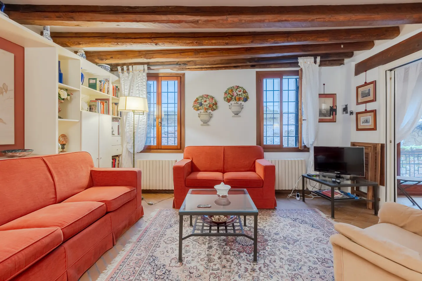 Living room with exposed wood beams, red sofas, a patterned rug, and a TV. Windows with white curtains let in natural light.
