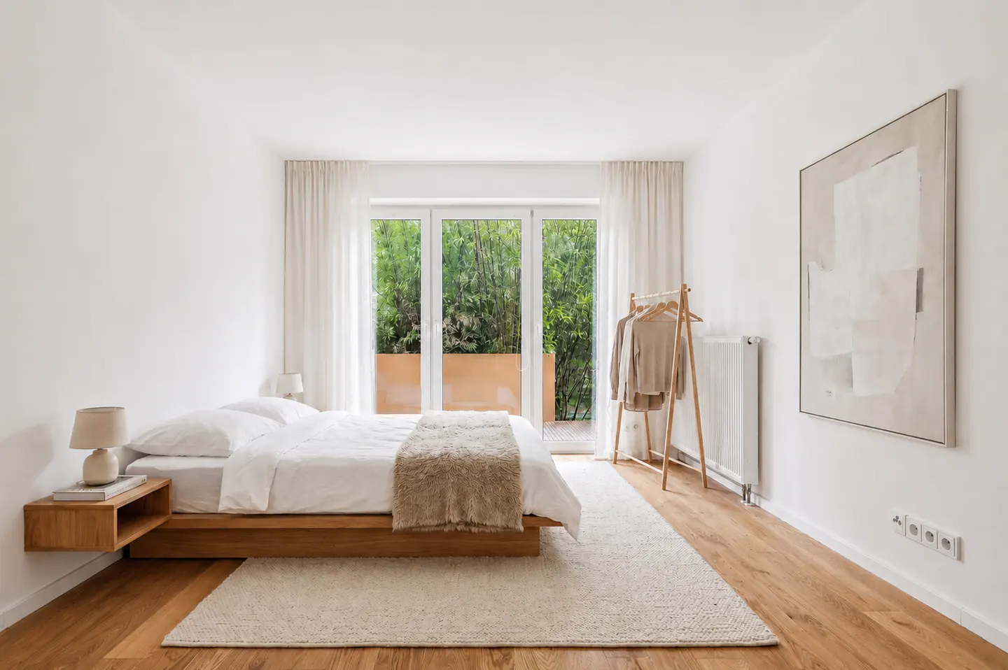 Minimalist bedroom with a wooden platform bed, white bedding, a beige throw, a clothing rack, and a large abstract painting.